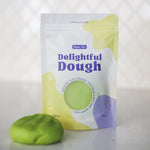 A pouch of Valia Co. Delightful Dough, soft play dough in green, shown next to a small ball of the same green dough on a light surface with a blurred white background.