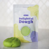 A pouch of Valia Co. Delightful Dough, soft play dough in green, shown next to a small ball of the same green dough on a light surface with a blurred white background.