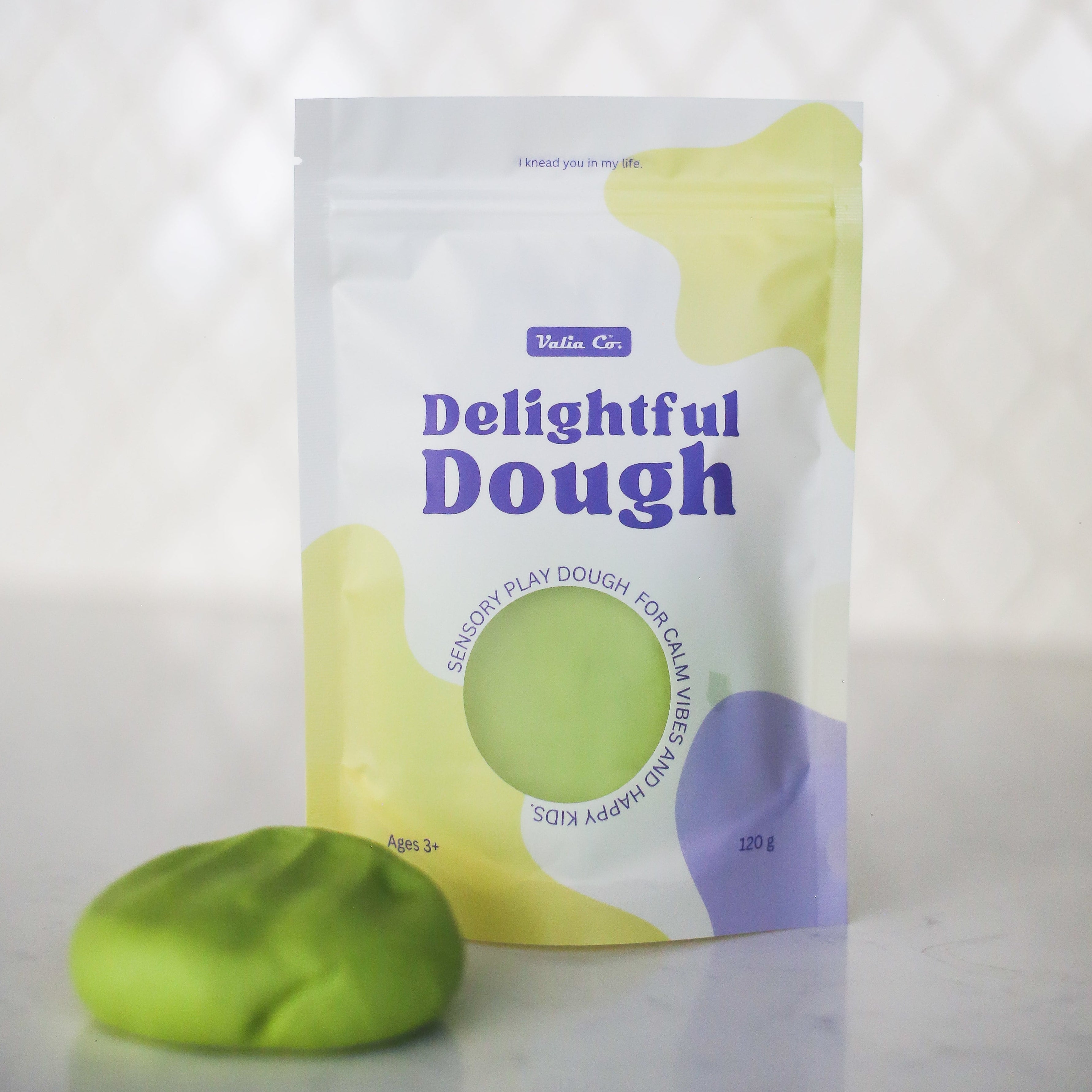 A pouch of Valia Co. Delightful Dough, soft play dough in green, shown next to a small ball of the same green dough on a light surface with a blurred white background.