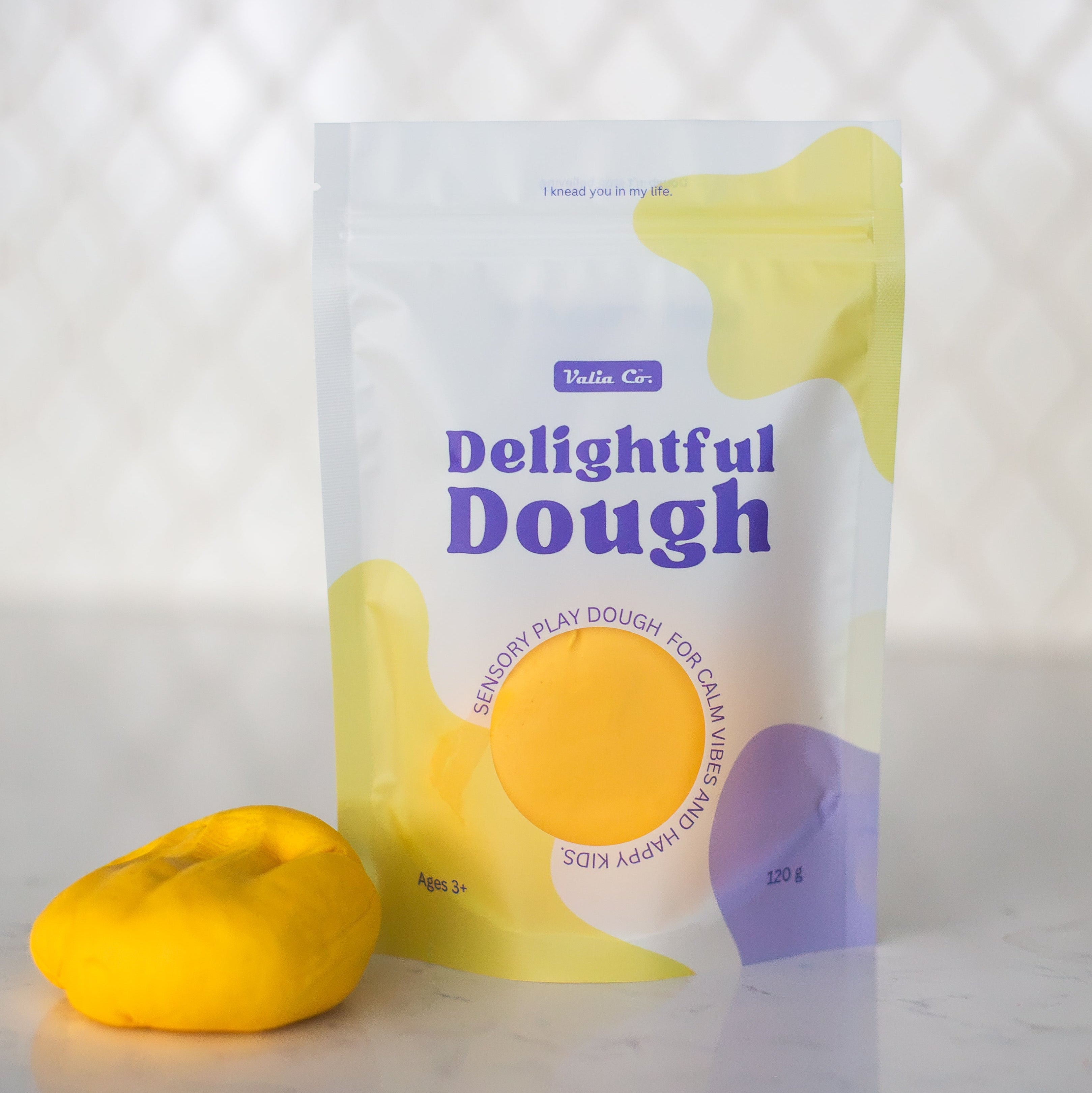 A stand-up pouch of Valia Co. Delightful Dough, play dough in yellow, shown on a light marble surface with a ball of yellow dough in front. The packaging features a white background with yellow and purple abstract shapes and blue text.