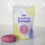 A stand-up pouch of Valia Co. Delightful Dough, play dough in pink, shown next to a small pink ball of dough on a light gray reflective surface with a textured gray background.