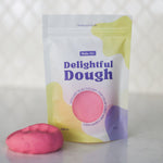 A pouch of Valia Co. Delightful Dough in pink, shown on a light countertop with a matching pink ball of dough in front of it against a tiled kitchen backsplash.