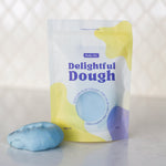 A stand-up pouch of Valia Co. Delightful Dough in a pastel blue color, shown next to a small, molded piece of the same blue dough on a light marble surface with a white tiled background.