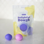 A stand-up pouch of Valia Co. Delightful Dough color-changing play dough shown on a light marble-like surface with two small dough balls, one purple and one pink, in front.