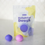 A stand-up pouch of Valia Co. Delightful Dough color-changing play dough shown on a light marble-like surface with two small dough balls, one purple and one pink, in front.