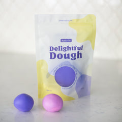 A stand-up pouch of Valia Co. Delightful Dough color-changing play dough shown on a light marble-like surface with two small dough balls, one purple and one pink, in front.