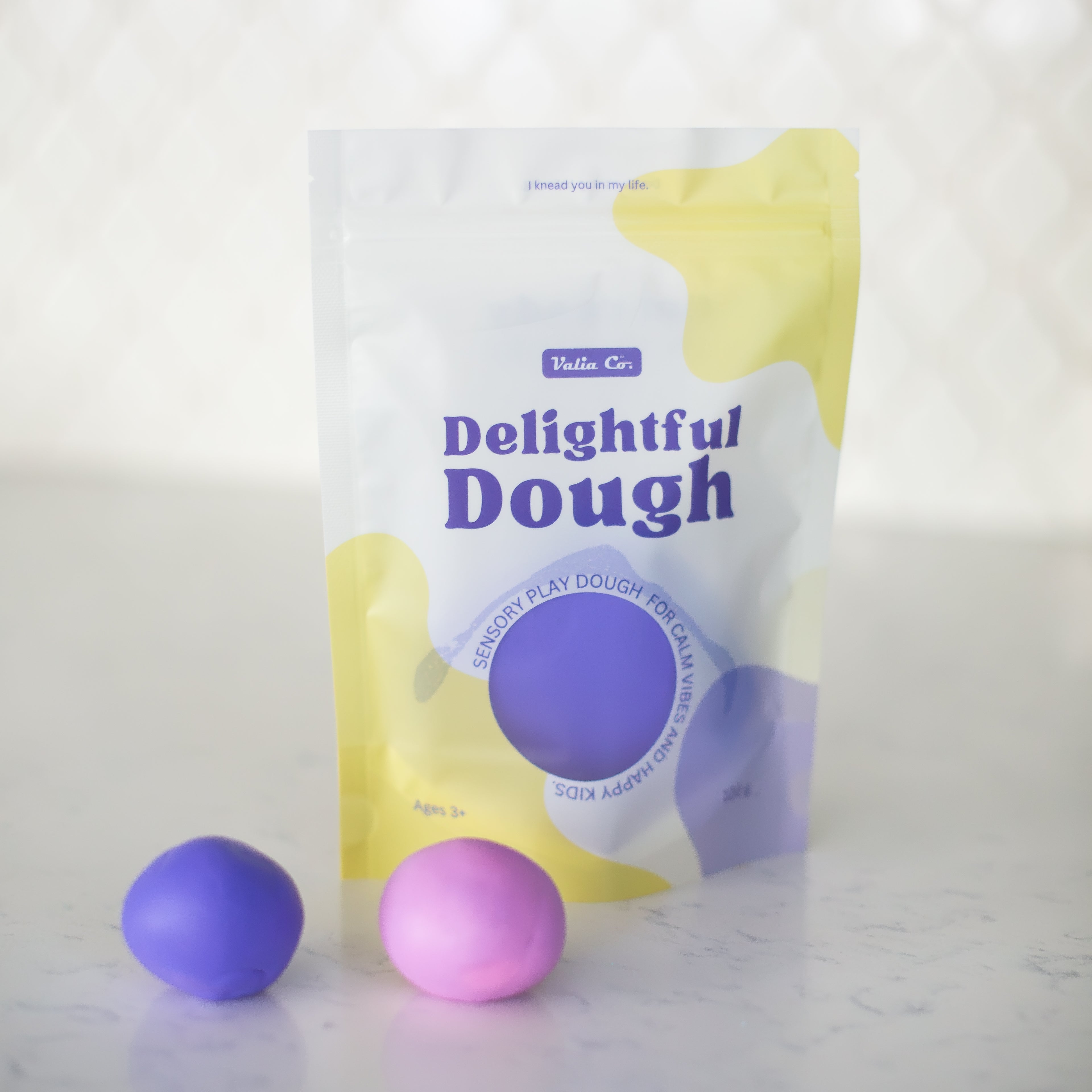 A stand-up pouch of Valia Co. Delightful Dough color-changing play dough shown on a light marble-like surface with two small dough balls, one purple and one pink, in front.