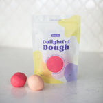 A stand-up pouch of Valia Co. color-changing Delightful Dough play dough stands on a light marble-like surface with two small balls of pink and peach-colored dough in front. 