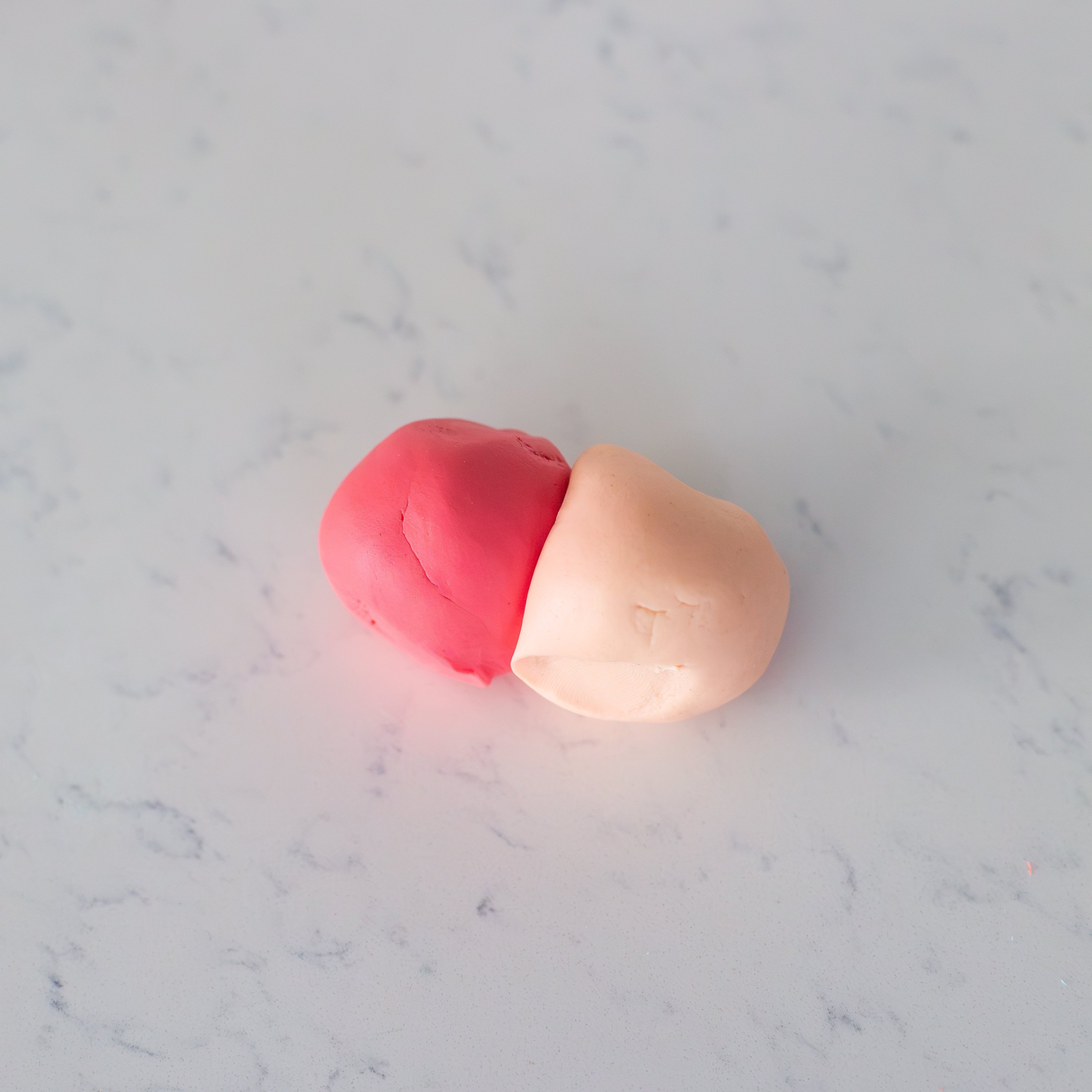 Two small pieces of clay or dough, one pink and one pale peach, resting on a light gray marble surface.