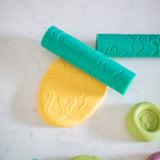 A close-up of colorful embosser stamps and a yellow piece of Valia Co. play dough stamped with a water snake design, resting on a white marble surface.